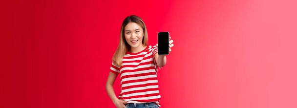 Check out my game score. Carefree good-looking blond asian woman extend arm showing smartphone screen smiling broadly stand relaxed introduce cool app, bragging bank account red background. - Foto, immagini
