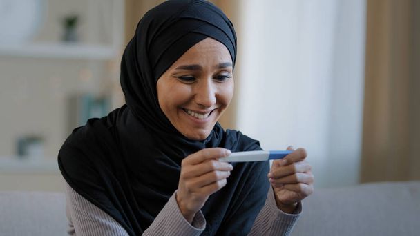 Portrét radostné překvapené dívka v hidžábu drží těhotenský test sedět v obývacím pokoji vzrušený s pozitivním výsledkem krásné islámské mladé ženy cítit radost po úspěšné léčbě plodnosti IVF koncept - Fotografie, Obrázek