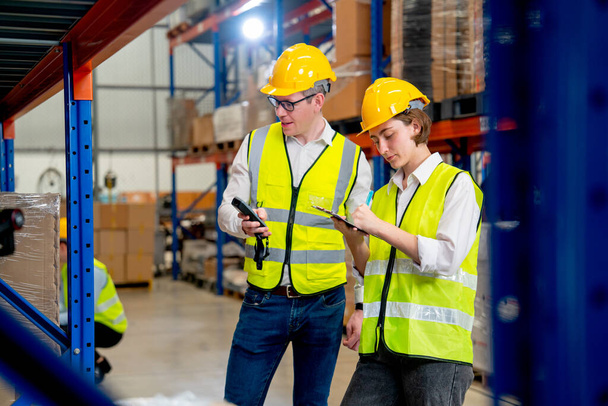 Warehouse worker man hold the barcode reader and discuss about the details of product on shelves with his co-worker woman and they ware safety protection in workplace. - Foto, imagen