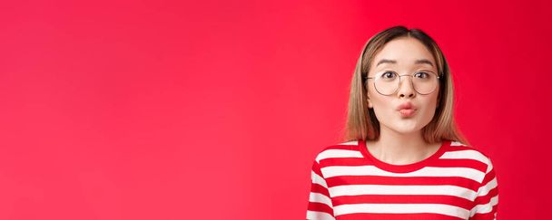 Cute pretty asian blond modern teenage girl folding lips anticipating tender romantic moment kiss, look camera wear glasses, blowing mwah, stand silly red background, feel passionate coquettish. - Photo, Image