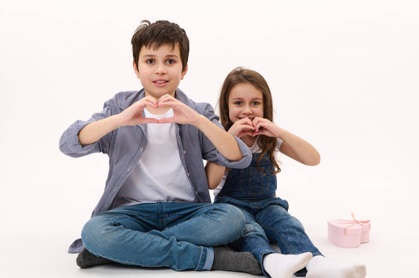 Rozkošné bělošské děti, chlapec a dívka v neformální košili a džínách džínách, ukazující srdce z prstů, usmívající se na kameru, izolované na bílém pozadí. Svatý Valentýn - Fotografie, Obrázek