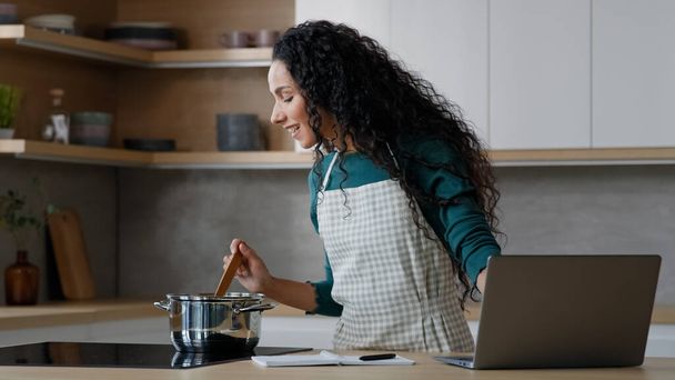 Jonge vrouwelijke chef-kok Arabisch spaanse vrouw kijken online tutorial culinaire video gebruik internet voedsel recept nuttig koken website op laptop koken dieetvoeding warme maaltijd gezonde voeding roeren smakelijke soep - Foto, afbeelding