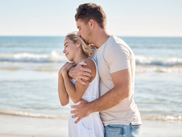 Пляж, счастливая пара, обнимающаяся с любовью, заботой и романтикой во время медового отпуска. Счастье, поцелуй и любящий мужчина обнимает свою жену сзади у океана на морском отдыхе на Мальдивах - Фото, изображение