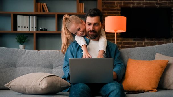 Cute active naughty girl noisy child plays with daddy hugs father distract parental attention disturbs parent from work on laptop at home single loving caucasian dad man calms down restless daughter - 写真・画像