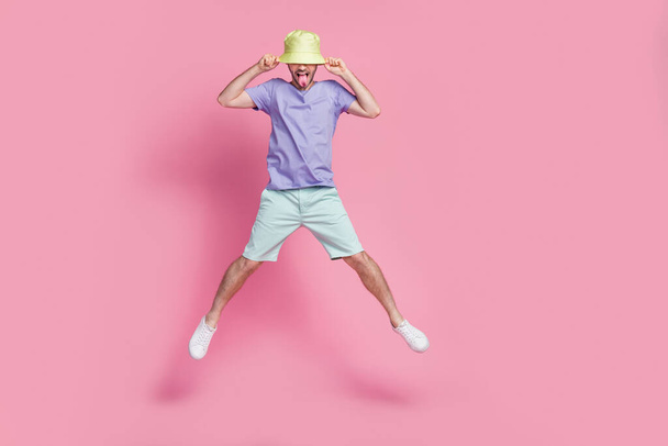 Full length photo of attractive young guy jump pull hat tongue out spread legs dressed trendy blue outfit isolated on pink color background. - Foto, immagini