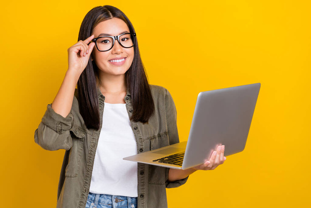 Porträt einer zufriedenen intelligenten Frau mit langer Frisur trägt eine khakifarbene Brille, die den Laptop isoliert auf gelbem Hintergrund hält. - Foto, Bild