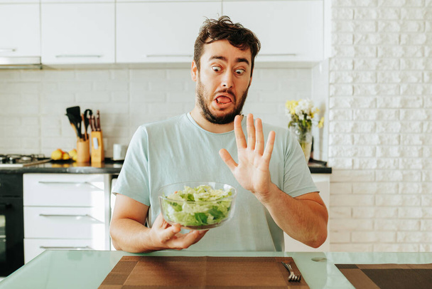young male guy sitting at a table in one hand holds bowl of salad with the other hand shows a stop, grimaces depicting a protest. want to be healthy, but I dont want to eat healthy food. Stop diet. - Fotoğraf, Görsel