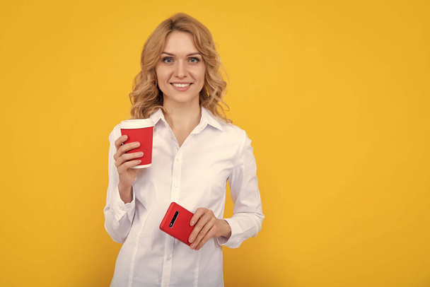 mujer rubia feliz con taza de café y teléfono inteligente sobre fondo amarillo. - Foto, imagen