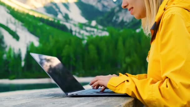 Женщина фрилансер типа текст на ноутбуке, сидя за деревянным столом против лесных гор и озера. Молодая женщина любит работать на свежем воздухе крупным планом - Кадры, видео