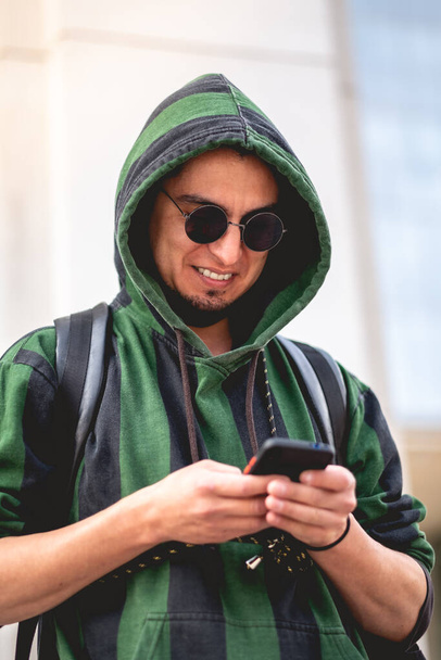 jong gelukkig guy met groene hoodie, ronde zonnebril en rugzak chatten in mobiele telefoon - Foto, afbeelding