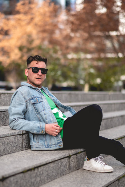 Νεαρός ξανθός τύπος μοντέλο με γυαλιά ηλίου, πράσινο t-shirt, τζιν σακάκι και μαύρο τζιν κάθεται σε πέτρινες σκάλες σε ένα πάρκο στην πόλη το απόγευμα - Φωτογραφία, εικόνα