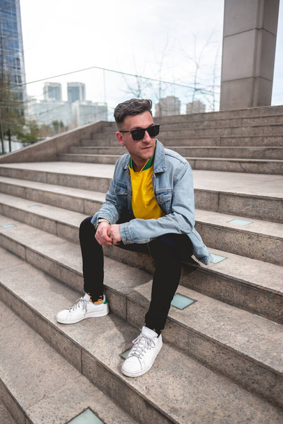 Mladý blonďatý modelář se slunečními brýlemi, žlutým tričkem, džínovou bundou a černými džínami sedí odpoledne na kamenných schodech v parku ve městě - Fotografie, Obrázek