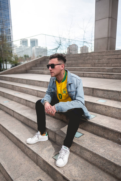 Mladý blonďatý modelář se čtyřlístkovým jetelem a slunečními brýlemi, žlutým tričkem, džínovou bundou a černými džínami sedí odpoledne na kamenných schodech v parku ve městě - Fotografie, Obrázek
