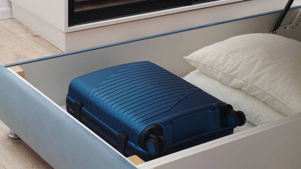 Storage drawer under bed with blue suitcase and white pillows indoors - 写真・画像