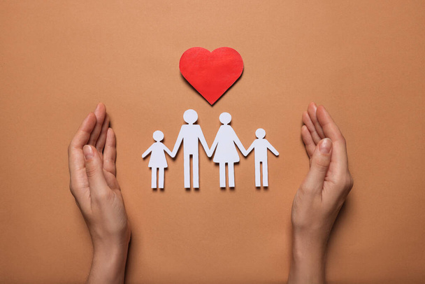 Woman protecting paper family figures and red heart on brown background, top view. Insurance concept - Фото, изображение
