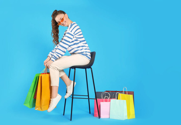 Happy woman in stylish sunglasses holding colorful shopping bags on stool against light blue background - Photo, image