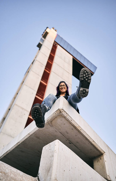 young latin woman wearing glasses blue pants sitting smiling at camera with hand hand out calm and relaxed next to concrete building - Urban concept - Foto, Bild