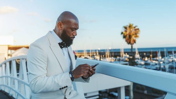 A profile view of a dashing bald African American man with a well-groomed black beard, in a fashionable white suit, texting using his smartphone while standing on a bridge next to a sailboats jetty - Foto, Imagem