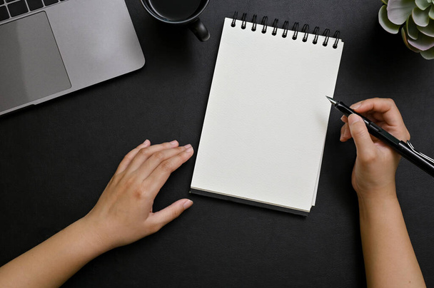 Πάνω άποψη, ένα γυναικείο χέρι κρατώντας στυλό, κρατώντας σημειώσεις σχετικά με σπιράλ σημειωματάριο στο σύγχρονο μαύρο γραφείο της. άδειο σημειωματάριο, laptop και διακόσμηση σε μαύρο φόντο. - Φωτογραφία, εικόνα