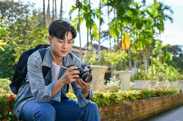 Guapo y feliz joven asiático viajero masculino descansando en un banco, revisando fotos en su cámara, viajando en sus vacaciones. - Foto, Imagen