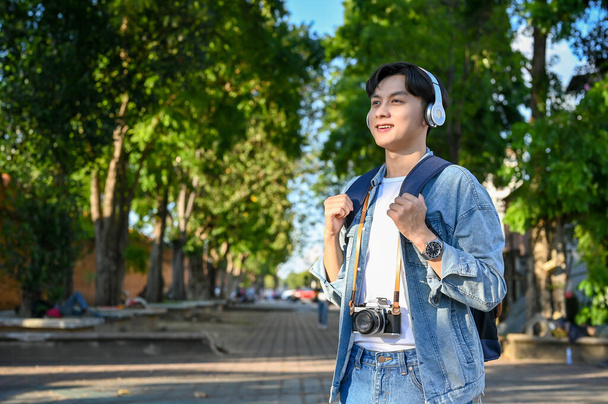 Hipster and handsome young Asian male traveler with his backpack, wearing headphones, enjoys strolling around the old city of Chiangmai, Thailand. Asia traveling concept - Photo, Image