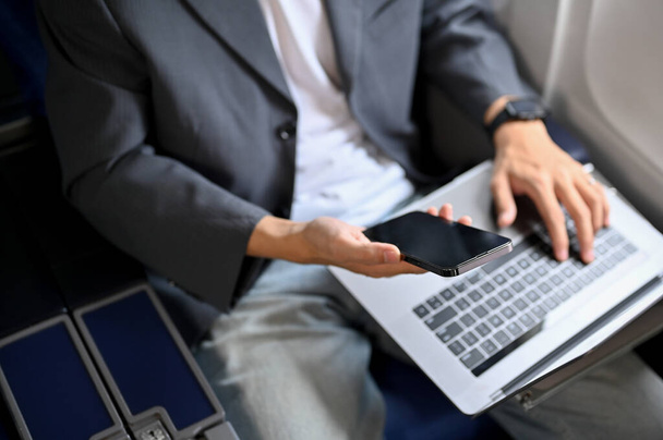 Над просмотром, занятой бизнесмен, использующий ноутбук и смартфон, работает над своими бизнес-задачами во время полета. - Фото, изображение