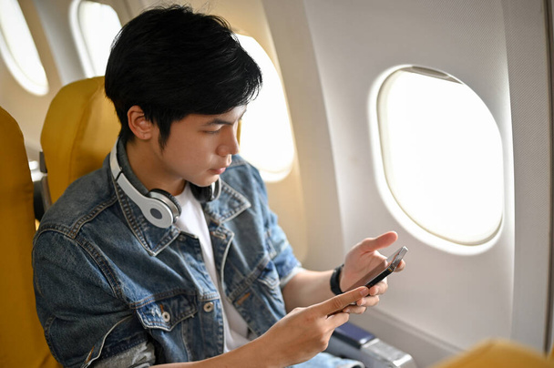 Handsome young Asian male passenger travel by airplane, sits at window seat in economy class using his smartphone after landing. Holiday journey concept - Photo, Image