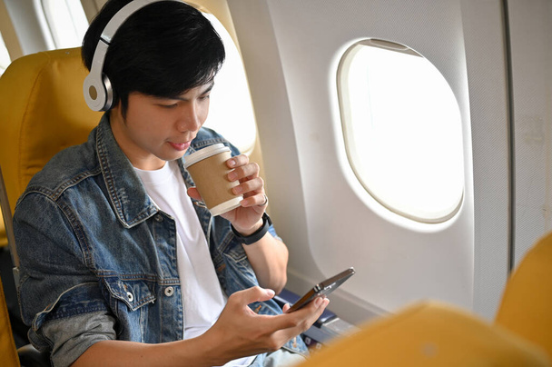 Komea aasialainen miesmatkustaja kuuntelee musiikkia kuulokkeidensa kautta, käyttää älypuhelinta ja siemailee kahvia lennon aikana määränpäähänsä. Kuljetusmatkan käsite - Valokuva, kuva