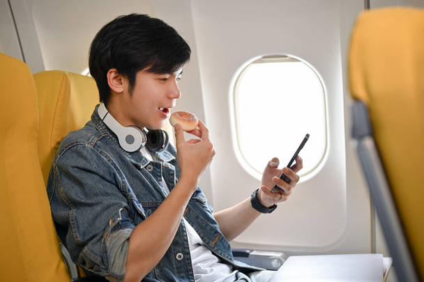 Happy and relaxed young Asian male passenger in an economy class, eating doughnut and using his smartphone during a flight. - Photo, Image