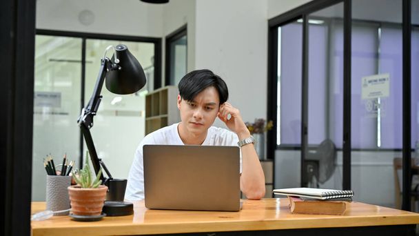 Όμορφος νεαρός Ασιάτης υπάλληλος γραφείου ή φοιτητής κολλεγίου, μπερδεμένος με την εργασία του, σκεπτόμενος και σχεδιάζοντας την εργασία του στο laptop. - Φωτογραφία, εικόνα