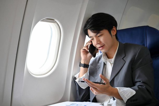 Επαγγελματίας και έξυπνος νεαρός Ασιάτης επιχειρηματίας με επίσημο κοστούμι που μιλάει στο τηλέφωνο, μοιράζοντας τις δουλειές του με τον συνεργάτη του κατά τη διάρκεια της πτήσης για το επαγγελματικό του ταξίδι. - Φωτογραφία, εικόνα