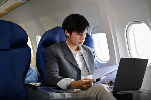Επαγγελματίας και επιτυχημένος νεαρός Ασιάτης επιχειρηματίας που εργάζεται, διαχειρίζεται τις επιχειρηματικές του εργασίες, προετοιμάζει έγγραφα συνάντησης κατά τη διάρκεια της πτήσης για το επαγγελματικό του ταξίδι. Έννοια των επιχειρήσεων και των μεταφορών - Φωτογραφία, εικόνα