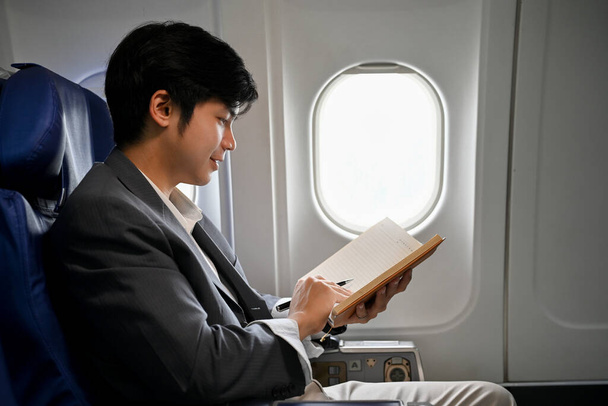 Όμορφος νεαρός Ασιάτης επιχειρηματίας ή διευθύνων σύμβουλος που διαβάζει ένα βιβλίο κατά τη διάρκεια της πτήσης για το επαγγελματικό του ταξίδι. Έννοια αεροπλάνου ή ιδιωτικού τζετ - Φωτογραφία, εικόνα