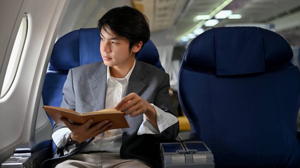 Joven empresario asiático exitoso o gerente ejecutivo masculino sosteniendo un libro mientras mira por la ventana, leyendo un libro durante el vuelo para su viaje de negocios. Concepto de avión o jet privado - Foto, Imagen