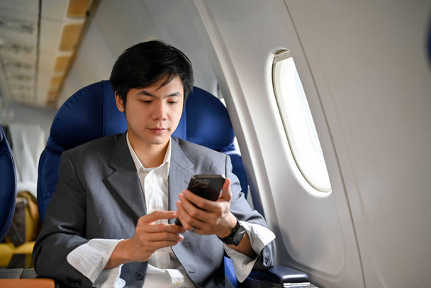 Smart und gut aussehender junger asiatischer Geschäftsmann mit seinem Smartphone während des Fluges im Privatjet zu seiner Geschäftsreise. - Foto, Bild