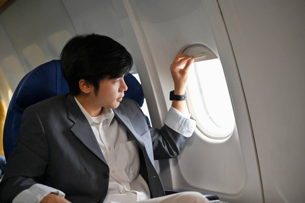 Schöner und smarter junger asiatischer Geschäftsmann oder Firmenchef im formellen Anzug sitzt am Fensterplatz in der Business Class, öffnet ein Fenster und blickt nach außen. - Foto, Bild