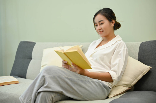 Relaksująca i spokojna Azjatka czyta książkę na przytulnej sofie w swoim salonie, Hobby i koncepcji czasu wolnego - Zdjęcie, obraz