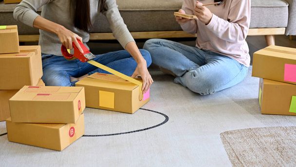 Zwei junge asiatische Inhaberinnen eines kleinen Online-Geschäfts halfen ihr, Bestellungen zu verpacken und ihre Pakete für den Versand in ihrem Wohnzimmer vorzubereiten. zugeschnittenes Bild - Foto, Bild