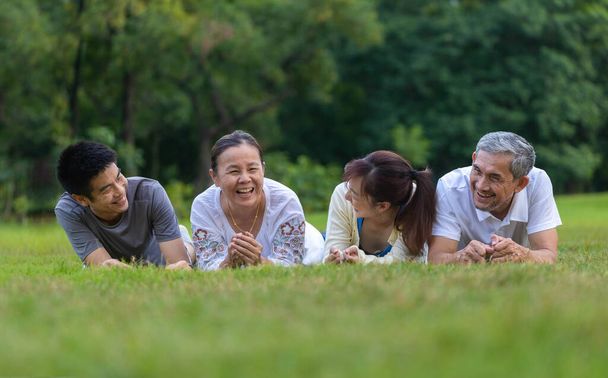 Група азіатських сімей з батьком, матір'ю, сином і дочкою лежить разом на трав'яному газоні і сміється в громадському парку під час вихідних діяльності для хорошого психічного здоров'я і відпочинку.  - Фото, зображення