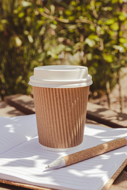 Eco recycling paper cup with coffee or tea on kraft paper with empty paper notebook on wooden bench. Concept of study work outdoors. Take away coffee to go. Copy space for text. Disposable Cardboard - 写真・画像