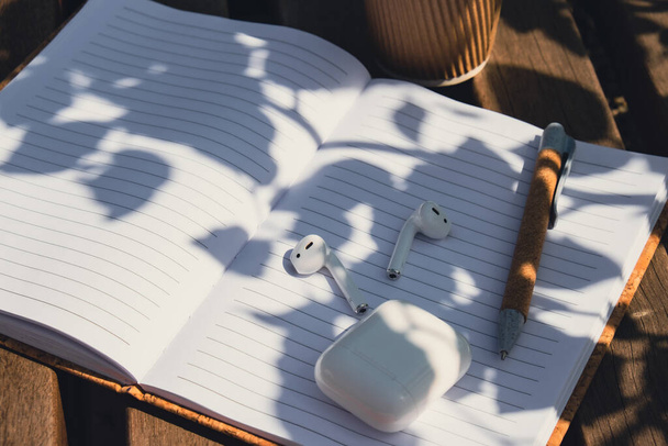 Kaffee zum Mitnehmen in Bastel-Recycling-Pappbecher mit Papier-Notizbuch mit drahtlosen Kopfhörern. Mockup Kaffeepause. Audio-Heilung, Klangtherapie Wellness-Rituale, positive psychische Gesundheit Hörgewohnheiten - Foto, Bild