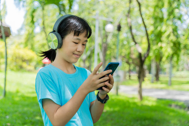Όμορφο κορίτσι του σχολείου με smartphone και ακούει μουσική με σύγχρονα ακουστικά ασύρματα στο πάρκο σε εξωτερικούς χώρους. - Φωτογραφία, εικόνα