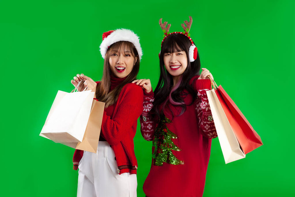 Young asian women long hairstyle lgbtq couple in red sweater in christmas theme clothes carrying paper shopping bags posing on green screen background. Merry Christmas. - Foto, imagen