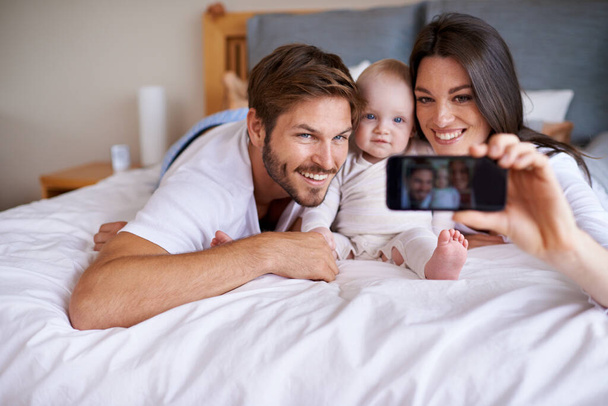 Семейное селфи. молодая пара лежит на кровати со своей малышкой и делает селфи со своим мобильным телефоном - Фото, изображение