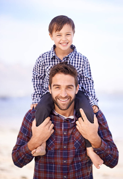 Μοιραζόμαστε ταιριαστά χαμόγελα απόλαυσης. Ένας πατέρας που κουβαλάει τον μικρό του γιο στους ώμους του με χαμόγελο. - Φωτογραφία, εικόνα