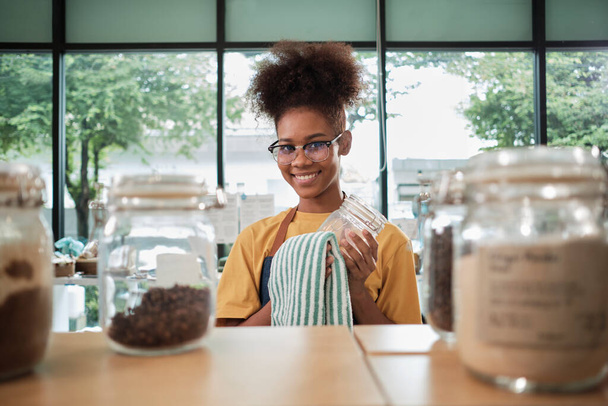 A young Black female shopkeeper cleans glass jars of natural organic products in reusable containers at a refill store, zero waste, a plastic-free grocery shop, and an eco-friendly retail business. - Фото, зображення