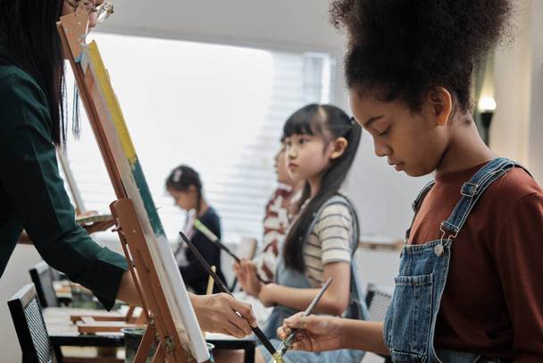 Afrikai amerikai lány összpontosít akril színes kép festés vászon diákokkal csoport művészeti osztályteremben, kreatív tanulás tehetségek és készségek általános iskolai stúdió oktatás. - Fotó, kép