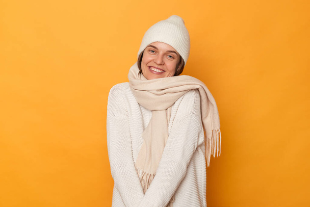 Indoor shot of shy cute woman wearing white jumper, cap and scarf posing against yellow wall, looking at camera with charming smile, expressing happiness, having high spirit, but hesitating. - Foto, Imagen