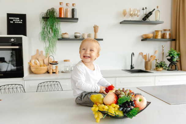 Un niño feliz sentado en la cocina sonriendo al lado de frutas y verduras.. Reductor de Comer Disfrutando de Alimentos a Base de Plantas - Foto, imagen