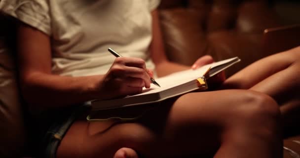 Un cuerpo femenino joven en el sofá con un diario. Una chica delgada escribe por la noche en un cuaderno, fondo marrón - Metraje, vídeo